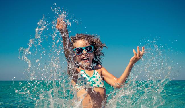 Bambina felice gioca spruzzando acqua al mare, sole e divertimento.