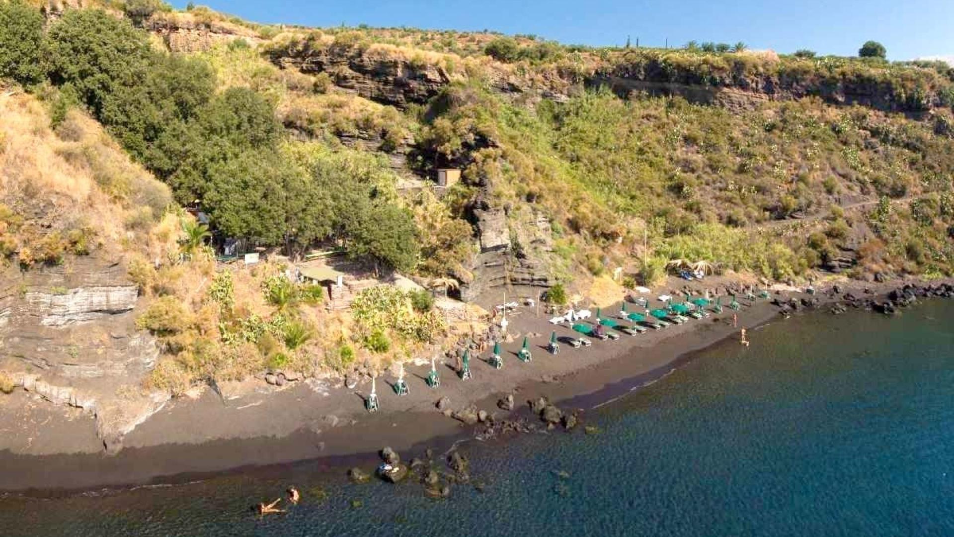 Spiaggia nera vulcanica, ombrelloni verdi, mare cristallino, scogliera verde, atmosfera tranquilla e naturale.