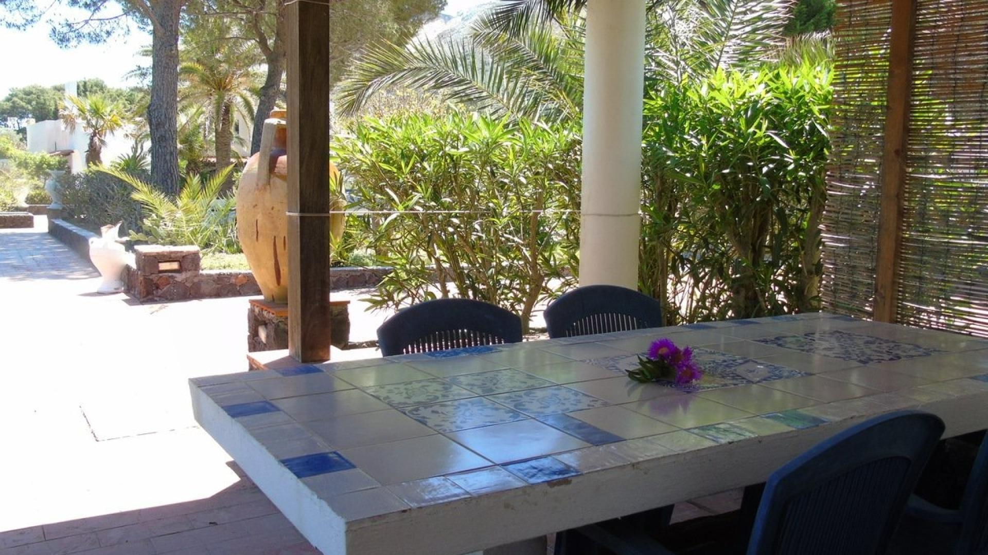 Veranda soleggiata con tavolo in ceramica, fiori, e giardino verdeggiante.