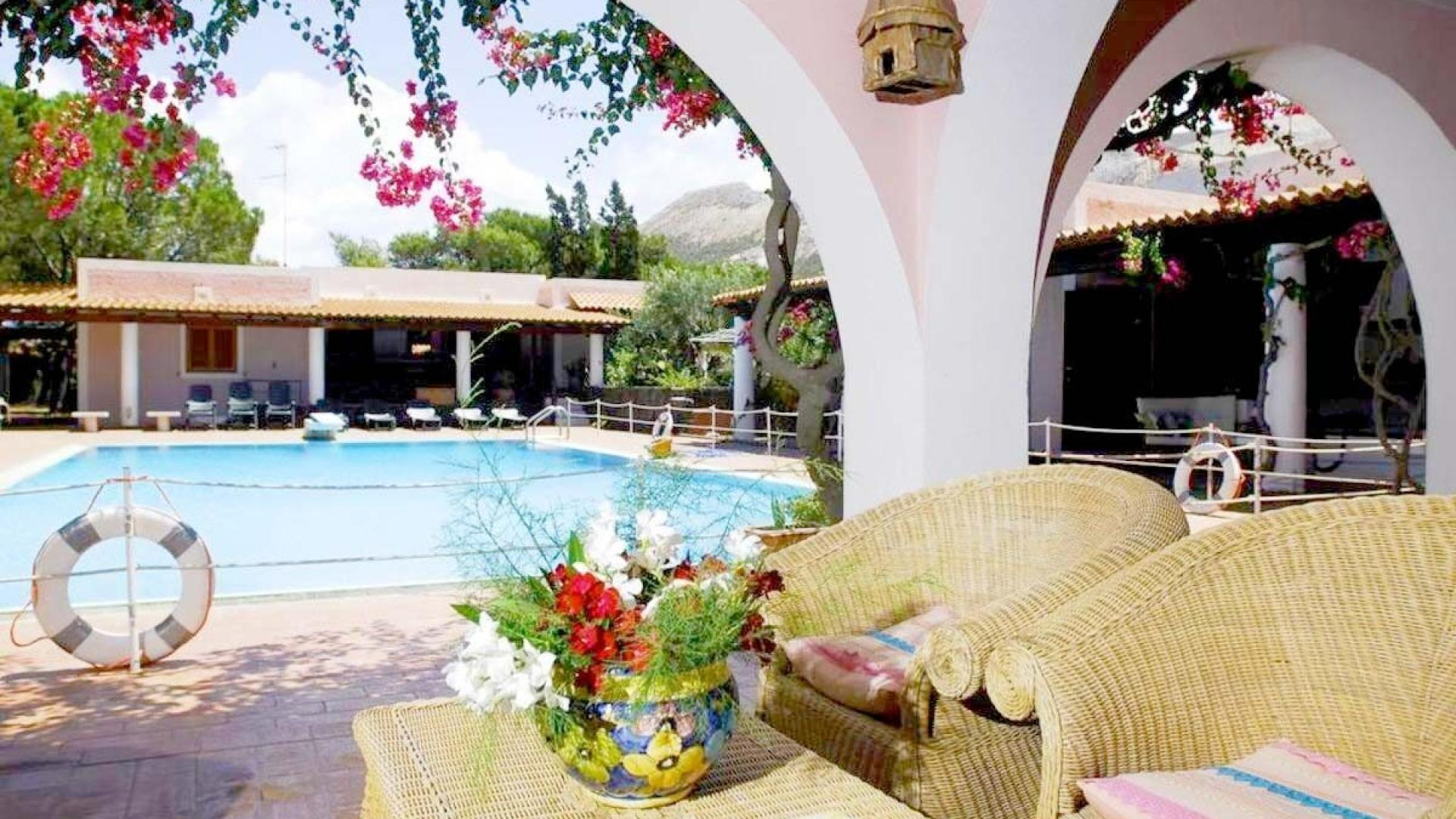Terrazza mediterranea con piscina, fiori colorati, sedie intrecciate, atmosfera rilassante e accogliente.