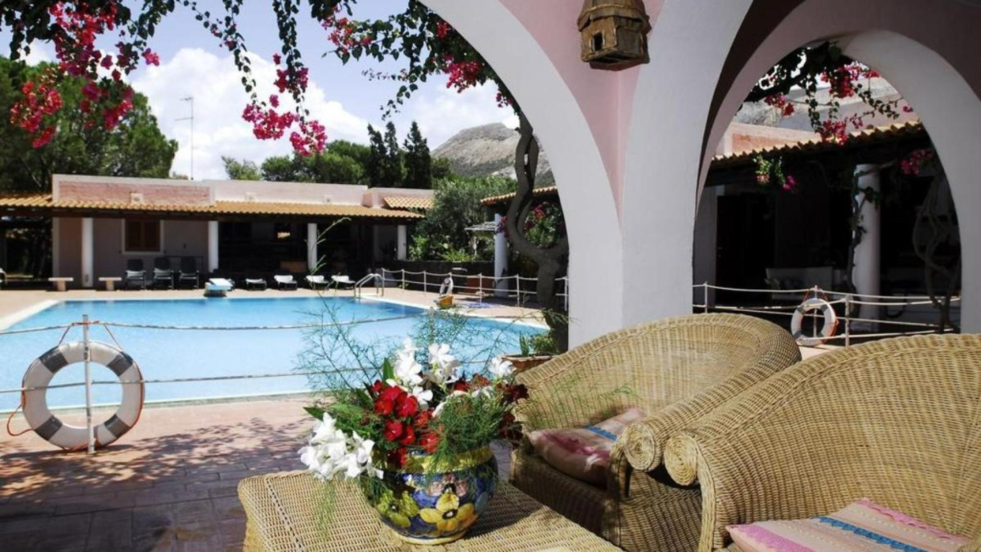 Giardino mediterraneo con piscina, archi fioriti, sedie in vimini e atmosfera rilassante.