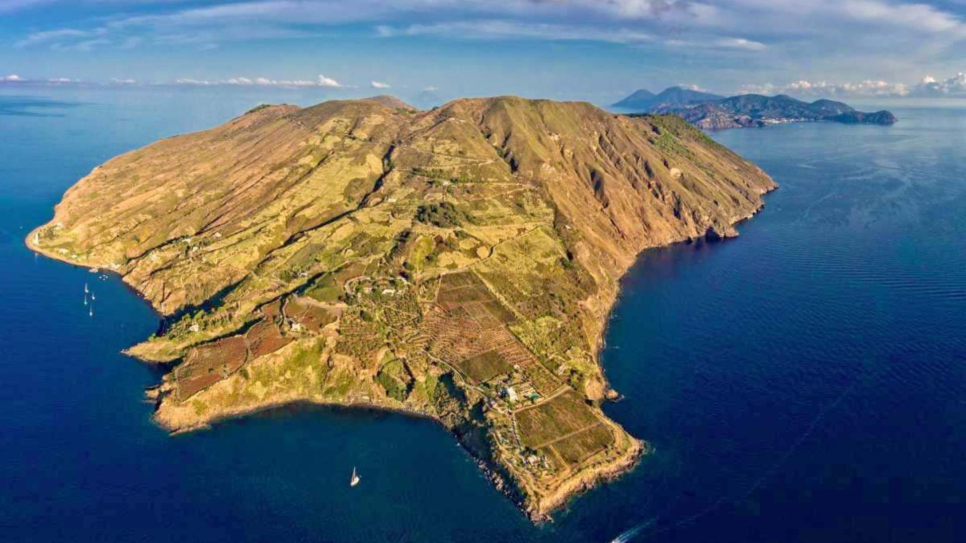 Isola verde e montuosa, circondata da mare blu, barche a vela e cieli sereni.