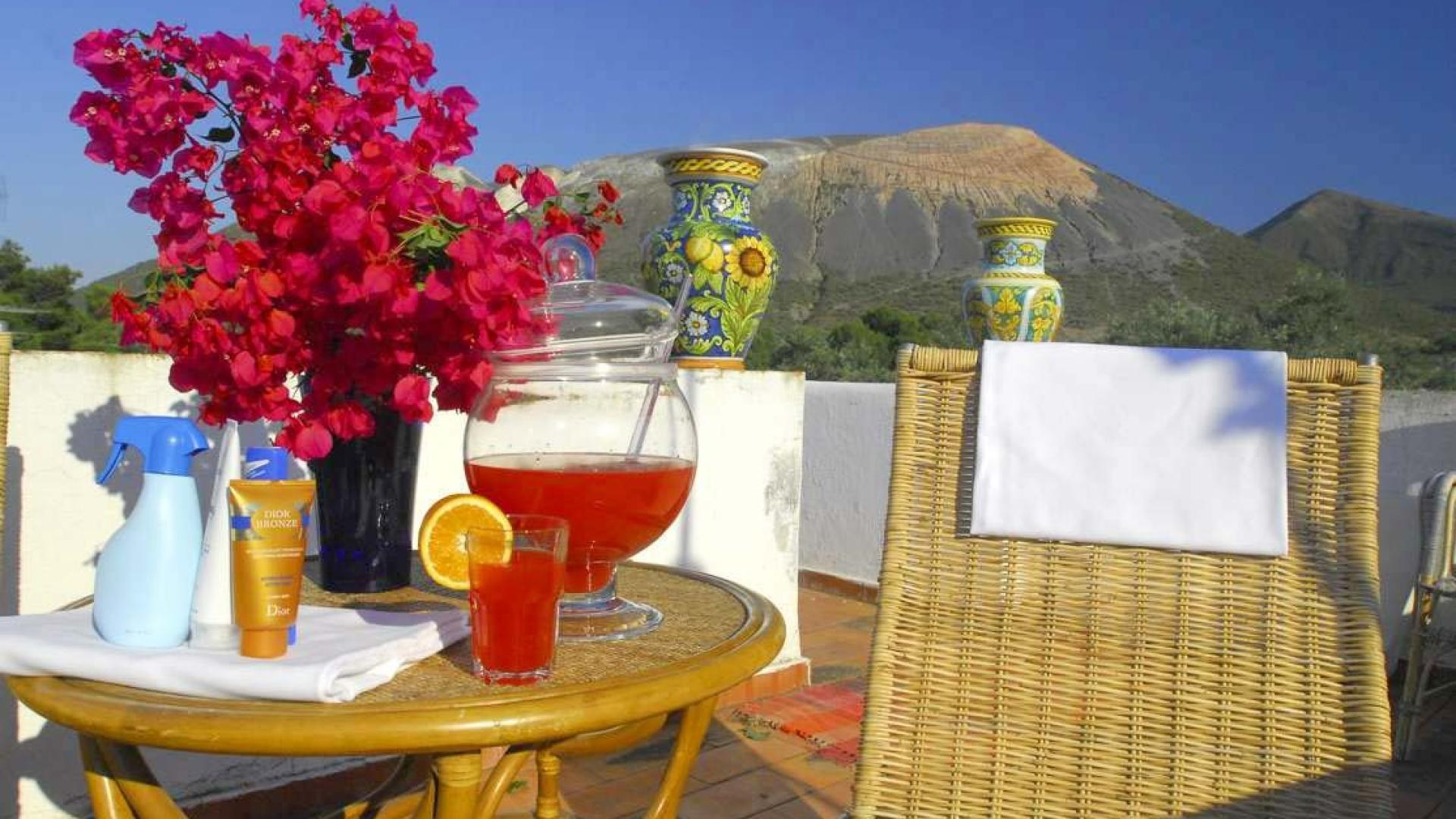 Terrazza soleggiata con fiori vivaci, bevanda fresca e montagne sullo sfondo.