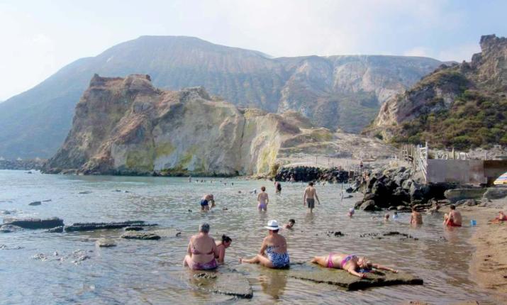 Spiaggia rocciosa, persone rilassate in acqua termale, montagne imponenti sullo sfondo.