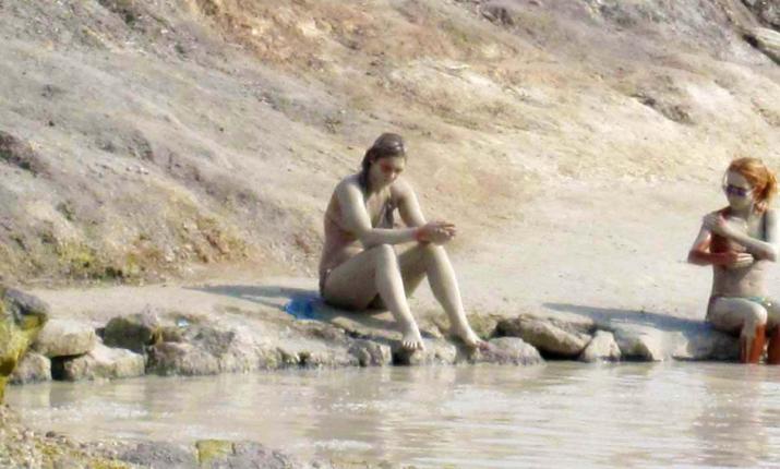 Due persone coperte di fango, sedute vicino a una pozza d'acqua naturale.