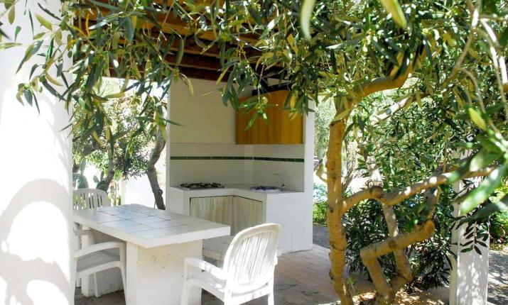 Cucina esterna immersa nel verde, tavolo bianco e sedie, atmosfera serena e naturale.