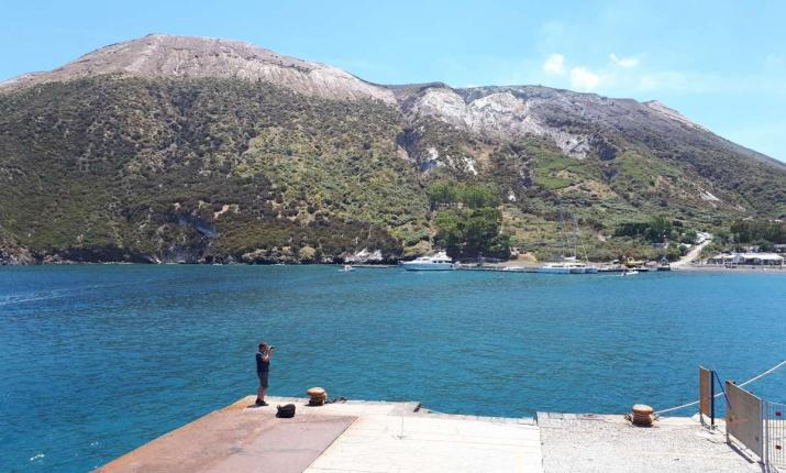 Una persona su un molo, di fronte a un'isola verde e mare blu intenso.