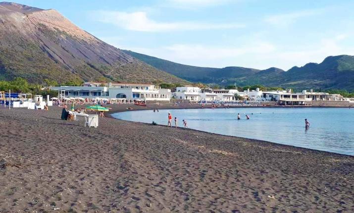 Spiaggia vulcanica nera, mare calmo, montagne imponenti, atmosfera rilassante e vacanziera.