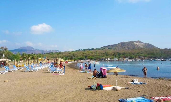 Spiaggia soleggiata, mare calmo, sdraio, ombrelloni colorati e colline verdi sullo sfondo.