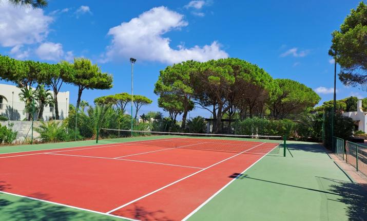 Campo da tennis soleggiato, circondato da pini e cielo azzurro, atmosfera serena e rilassante.