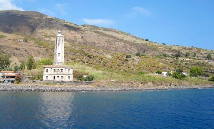 Faro solitario su costa verde, montagne sullo sfondo, cielo azzurro e mare calmo.