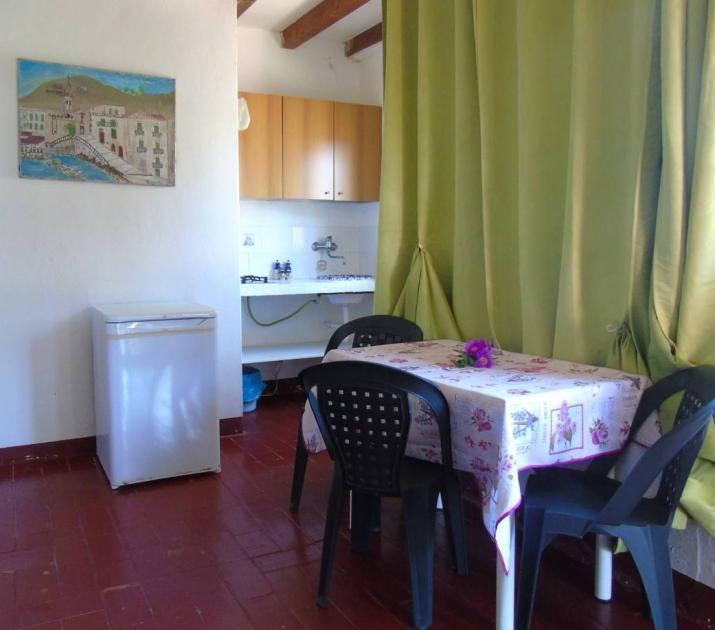 Cucina semplice con tavolo fiorito, sedie nere e tenda verde, atmosfera accogliente e luminosa.
