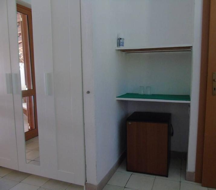 Stanza semplice, armadio bianco, mini frigo, bicchieri, porta chiusa, atmosfera tranquilla.