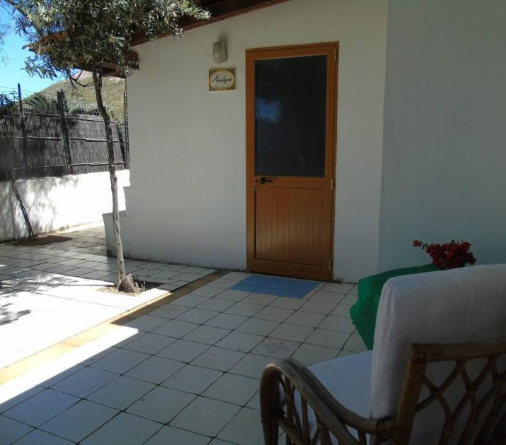 Cortile soleggiato con sedia in vimini, porta in legno, pianta e atmosfera serena.