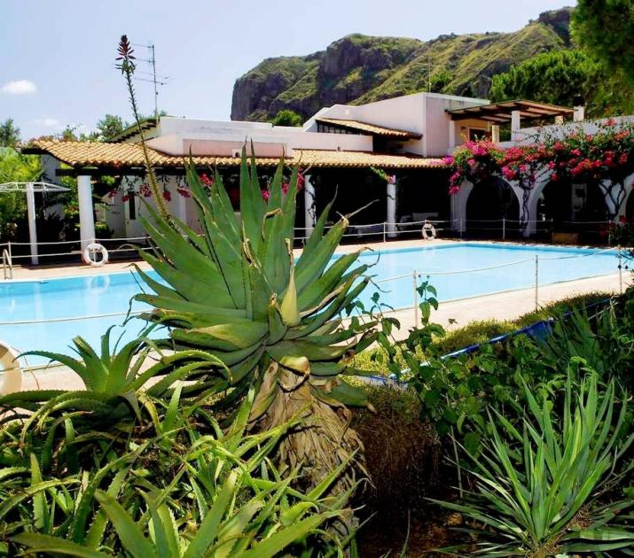 Piscina serena con piante lussureggianti, fiori colorati e montagne sullo sfondo.
