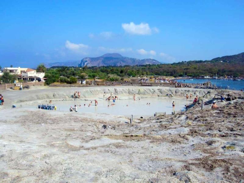Piscina naturale di fango, persone rilassate, paesaggio vulcanico e mare azzurro sullo sfondo.