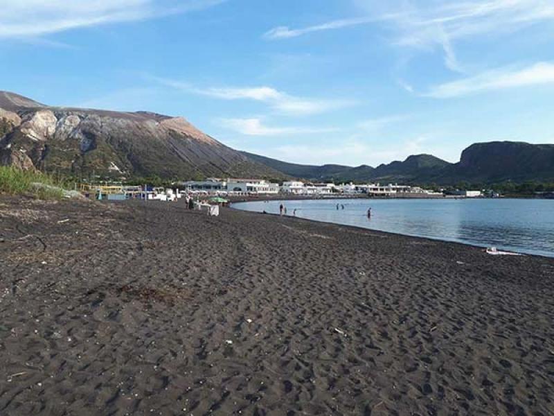 Spiaggia di sabbia nera, mare calmo, montagne imponenti sotto un cielo azzurro.